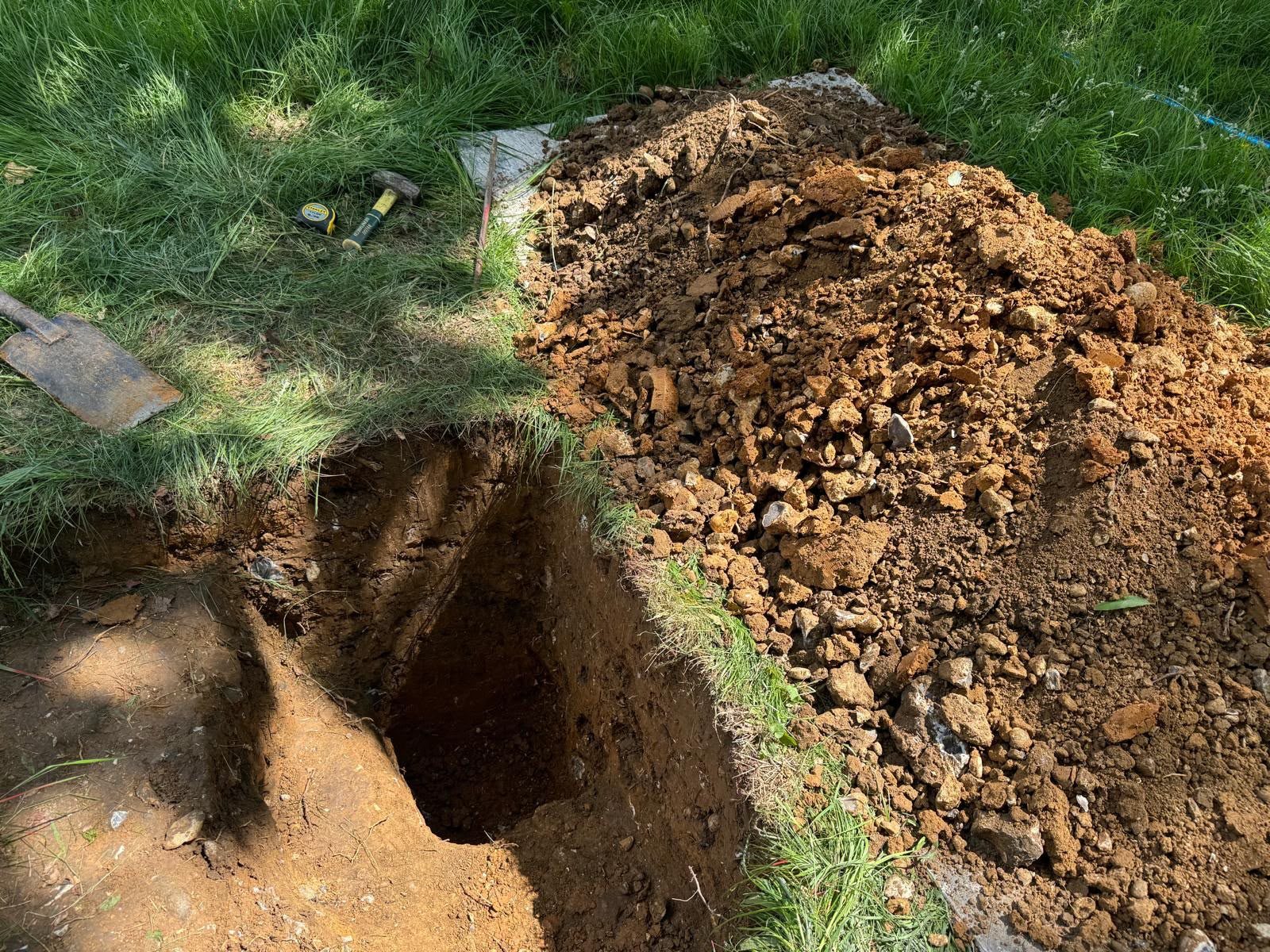 Hand-dug trial pit in a lawn with the excavated spoil, spade and tape measure alongside, ready for infiltration testing