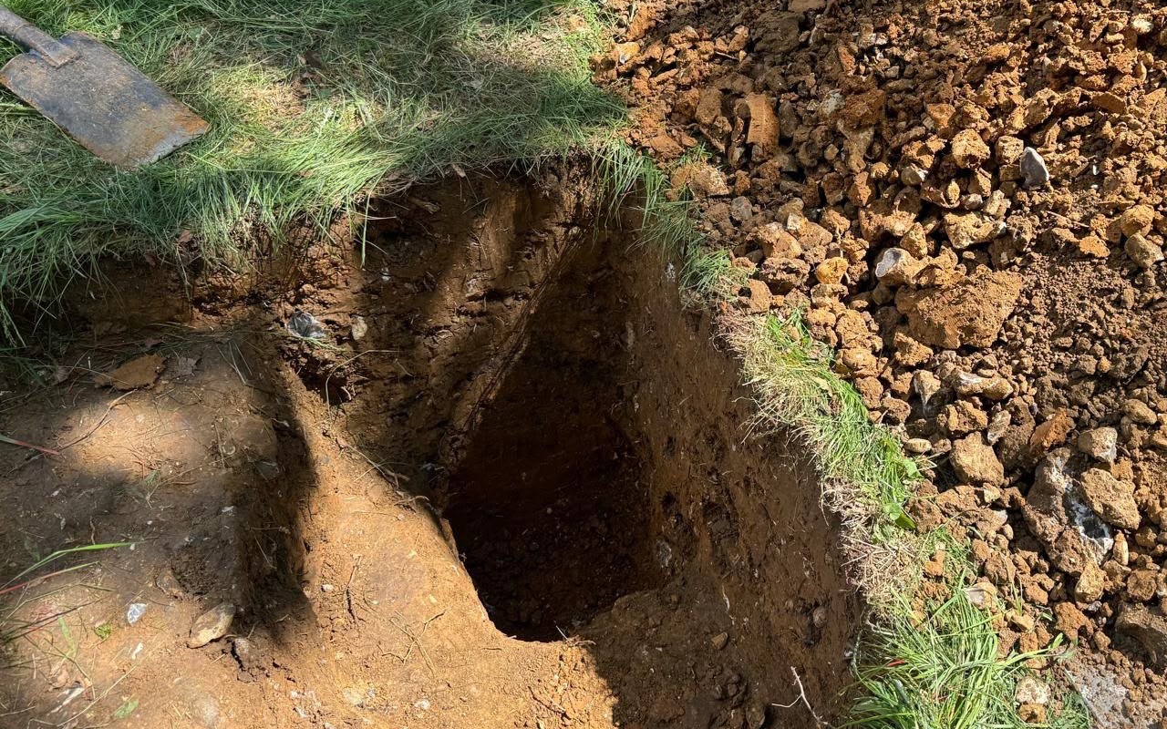 Close view of a hand-dug trial pit in a lawn, with the spade and excavated spoil alongside, during infiltration testing
