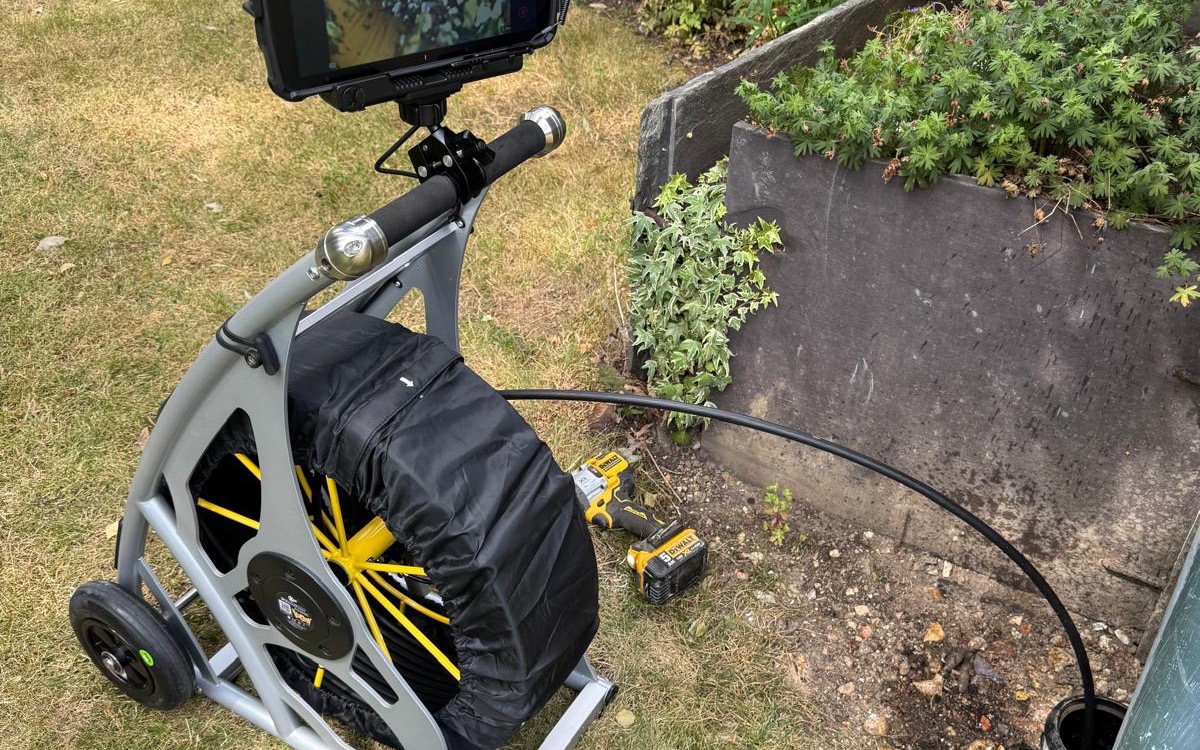 Flowcraft CCTV drain survey reel on a lawn, with the camera cable fed into a drain opening beside a raised garden bed